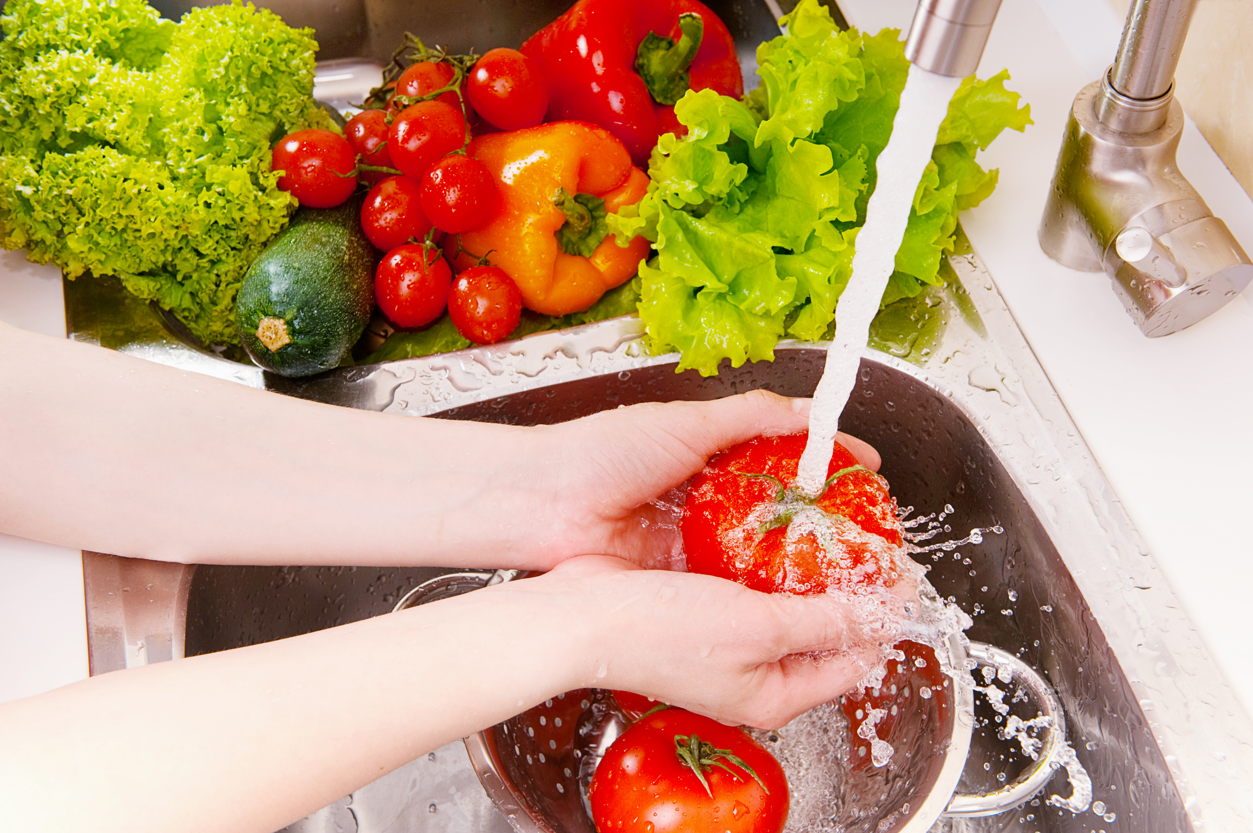 A pair of hands holds a tomato under running water, with lettuce, cucumber, peppers and cherry tomatoes piled on the counter next to the sink.