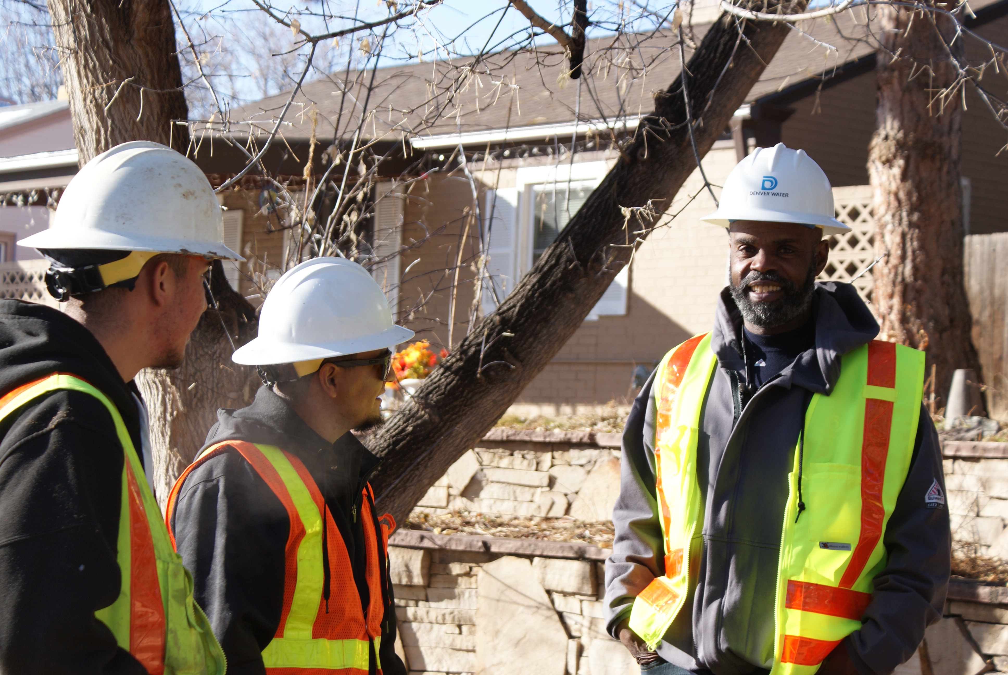 Three men in Denver Water hard hats and bright yellow and orange safety vests talk in front of a house.
