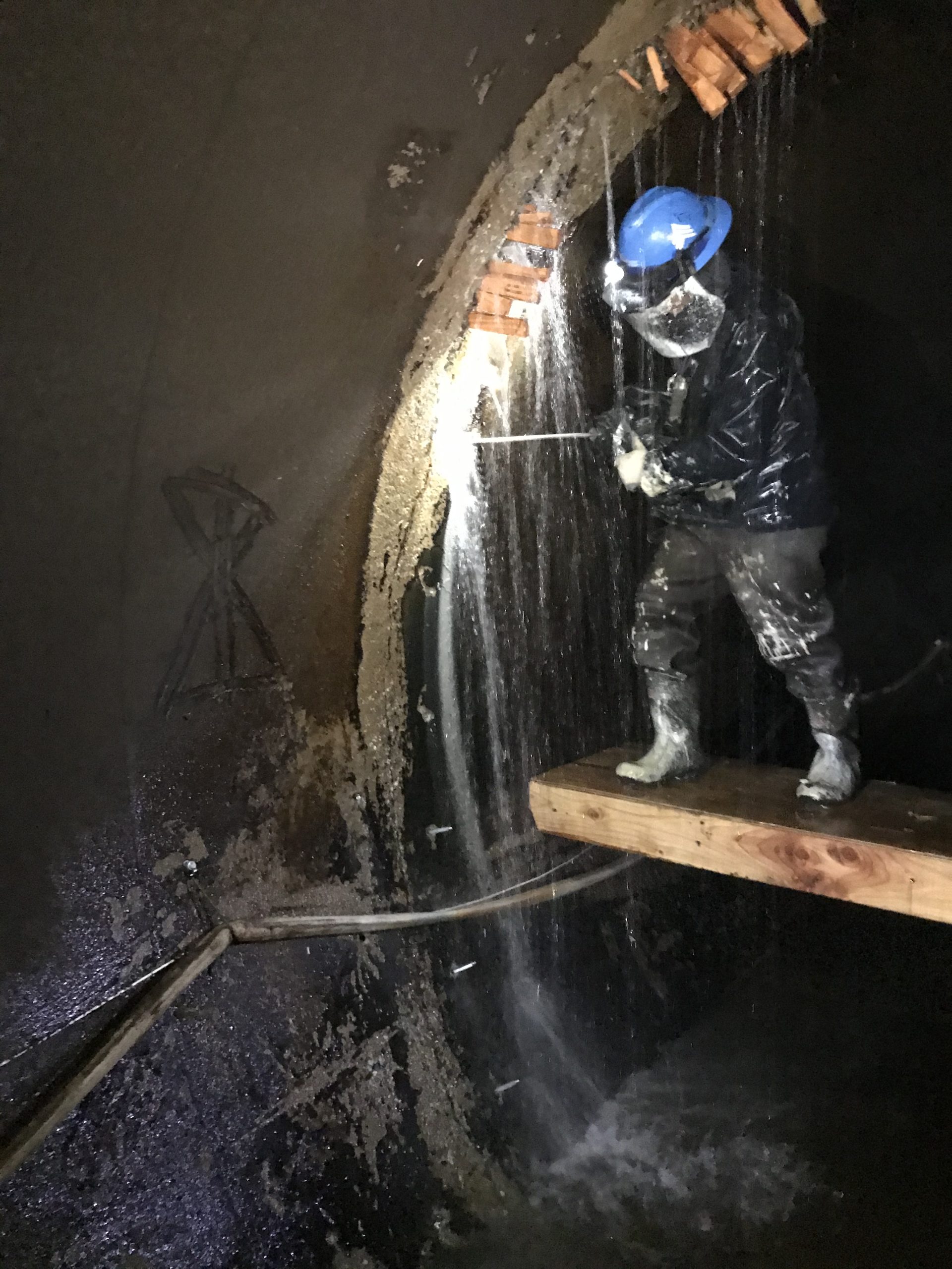 A man stands on a platform, in a hard hat and safety gear, spraing something onto a wall of a tunnel.