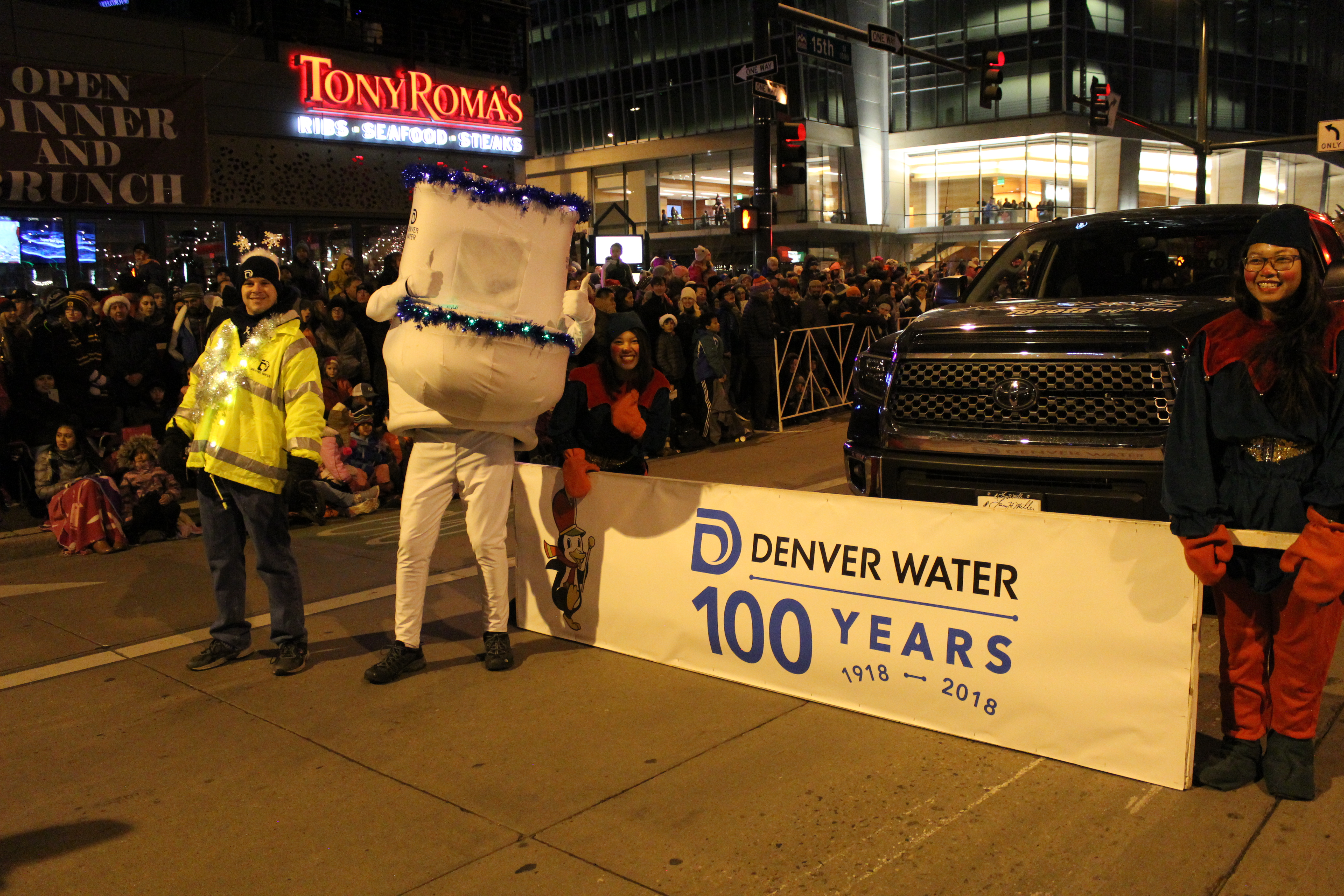 An image of a Denver Water volunteer in a yellow jacket, the running toilet giving a thumbs-up sign, and the Denver Water banner held by volunteers in elf costumes.