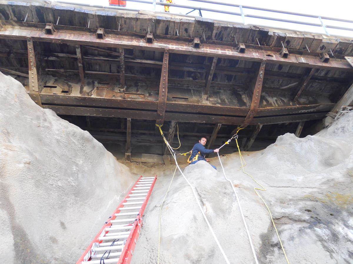 A ladder leans against a slab of rock and overhead is the underside of a giant bridge, with beams and other infrastructure dwarfing a man, roped in for safety, doing an inspection.