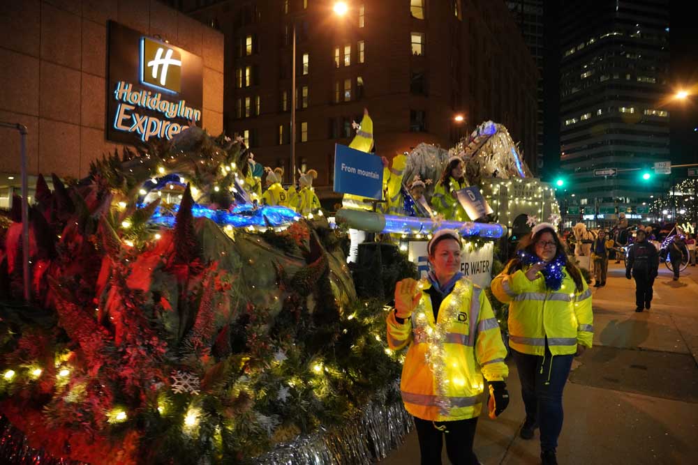 Two women in yellow Denver Water safety jackets garland and strings of lights, walk down a street.